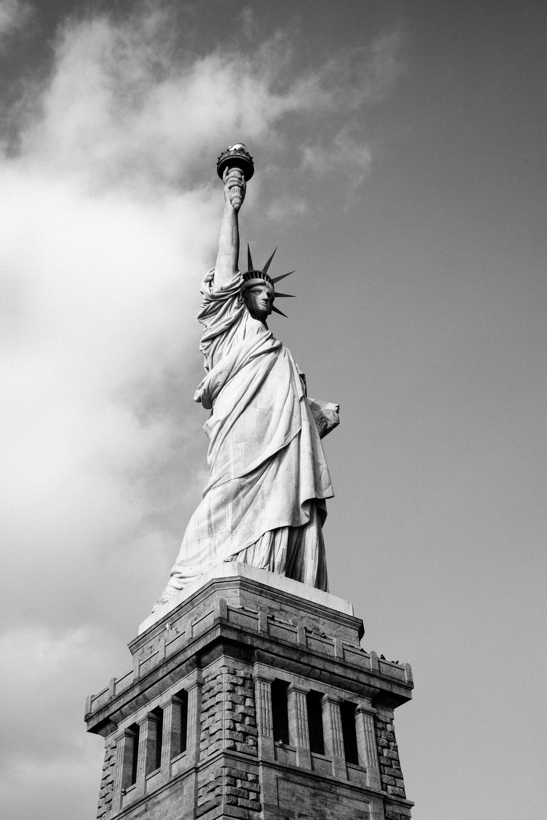 The Statue of Liberty against a clouded sky, torch raised. Black and white.